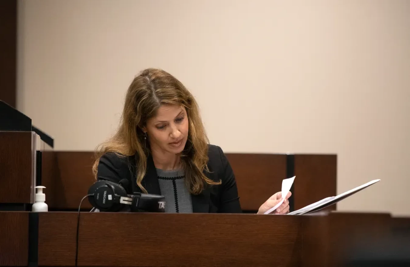 A woman with a dark suit sits on the witness stand, flipping through a folder